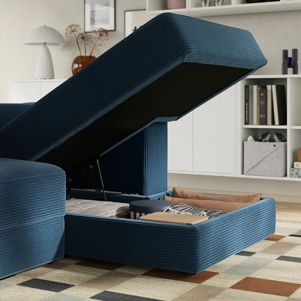 A blue sectional sofa with raised storage, hiding a mattress, pillows, and blanket. The sofa includes footrest and has a cosy, inviting look.