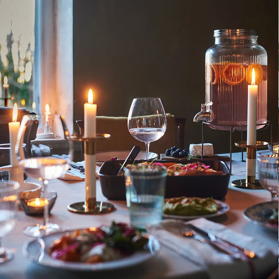 A dinner table set with plates, glasses, candles, and a VARDAGEN jar stand in black, holding a jar with a tap, surrounded by hanging lamps.