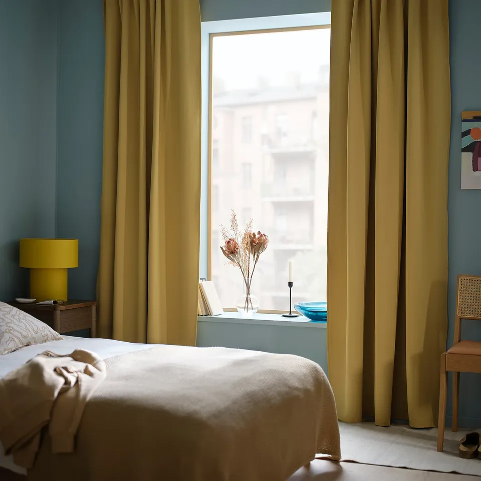 Bedroom with MAJGULL room darkening curtains in beige-yellow, matching bedding, and wooden furniture, featuring a window with a city view.