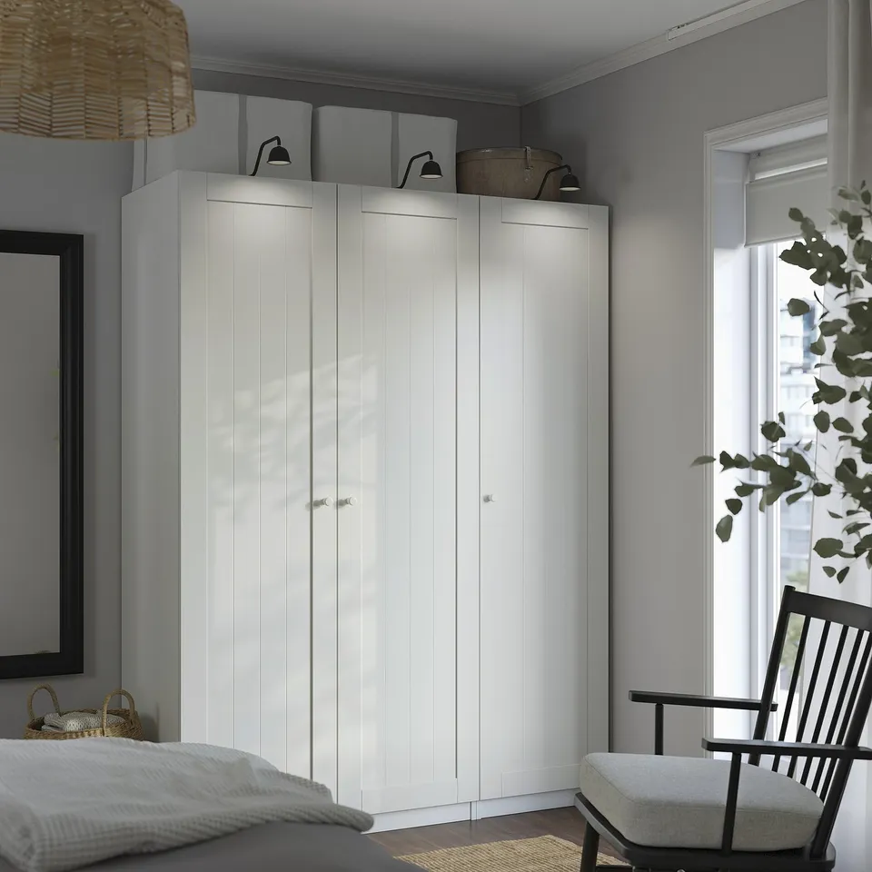 Large white PAX wardrobe, tall and slim, dominating room with grey walls. Topped with wicker basket and lamps, beside black rocking chair and window.