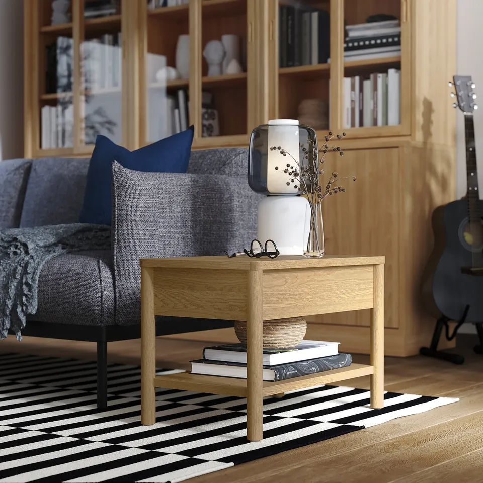 Wooden side table with drawer, light colour, black and white striped rug, grey couch, books, vase with dried flowers, guitar.