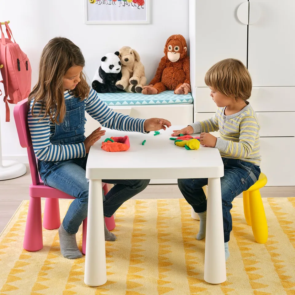 Two children play with colourful modelling dough at a small table, seated on bright chairs. Stuffed animals sit on a nearby bench.