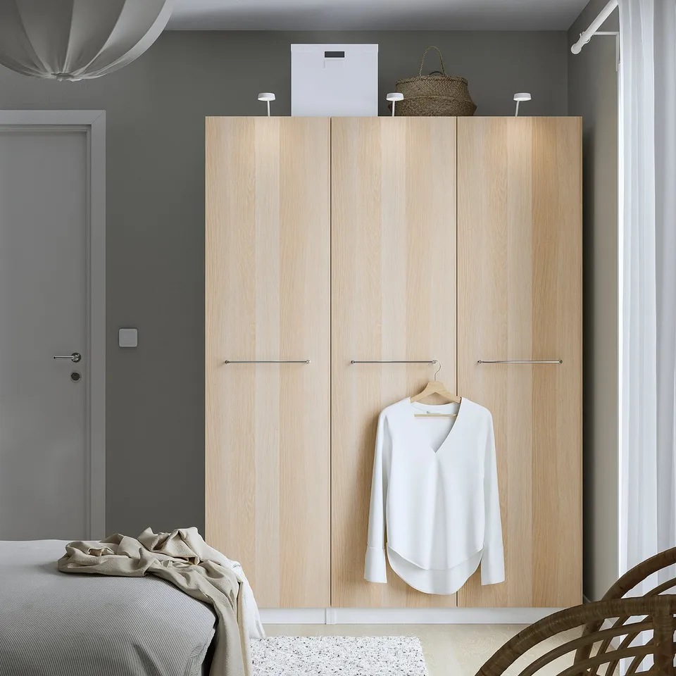 Minimalist bedroom with large light wooden PAX closet, white blouse hanging, woven bag atop, beside beige bed.