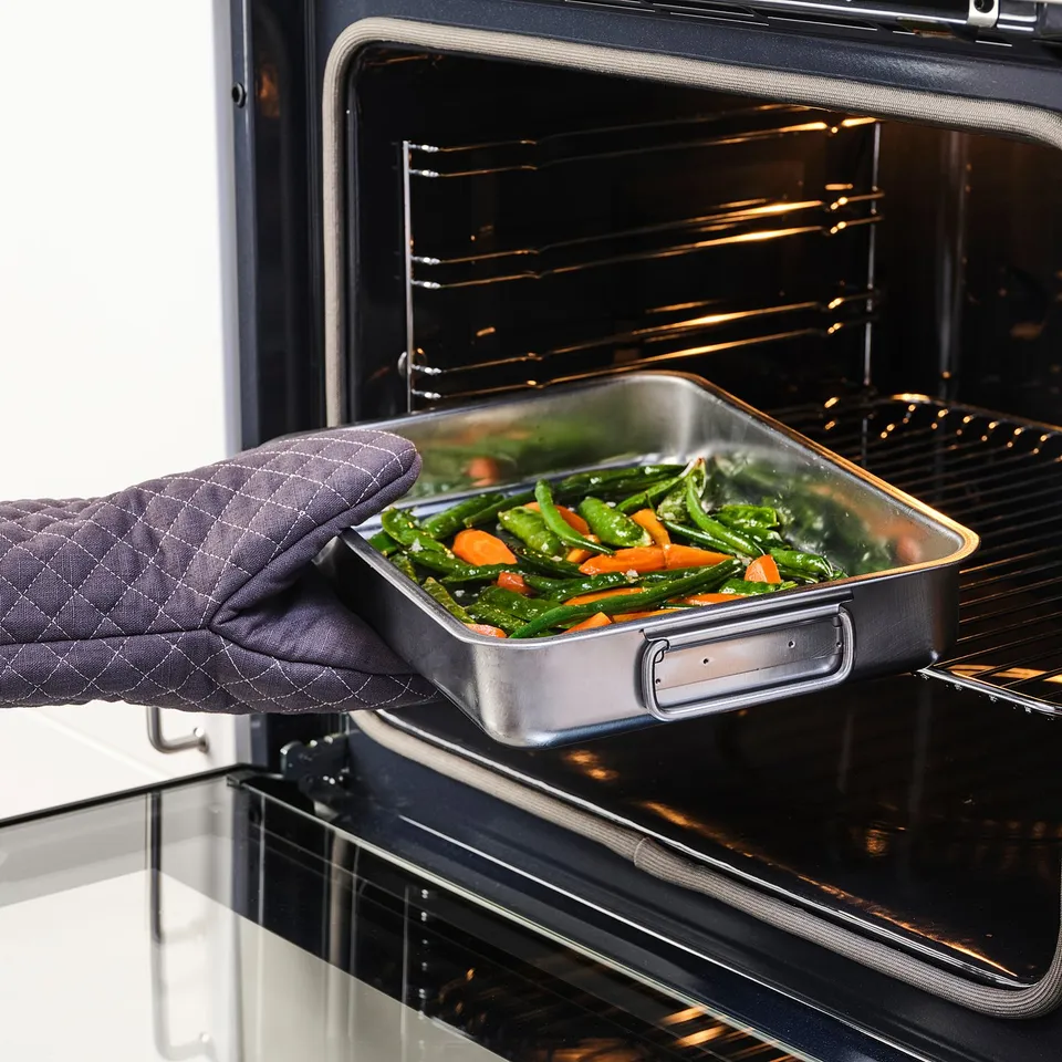 Person with oven mitt removes baking tray filled with roasted veggies from oven.