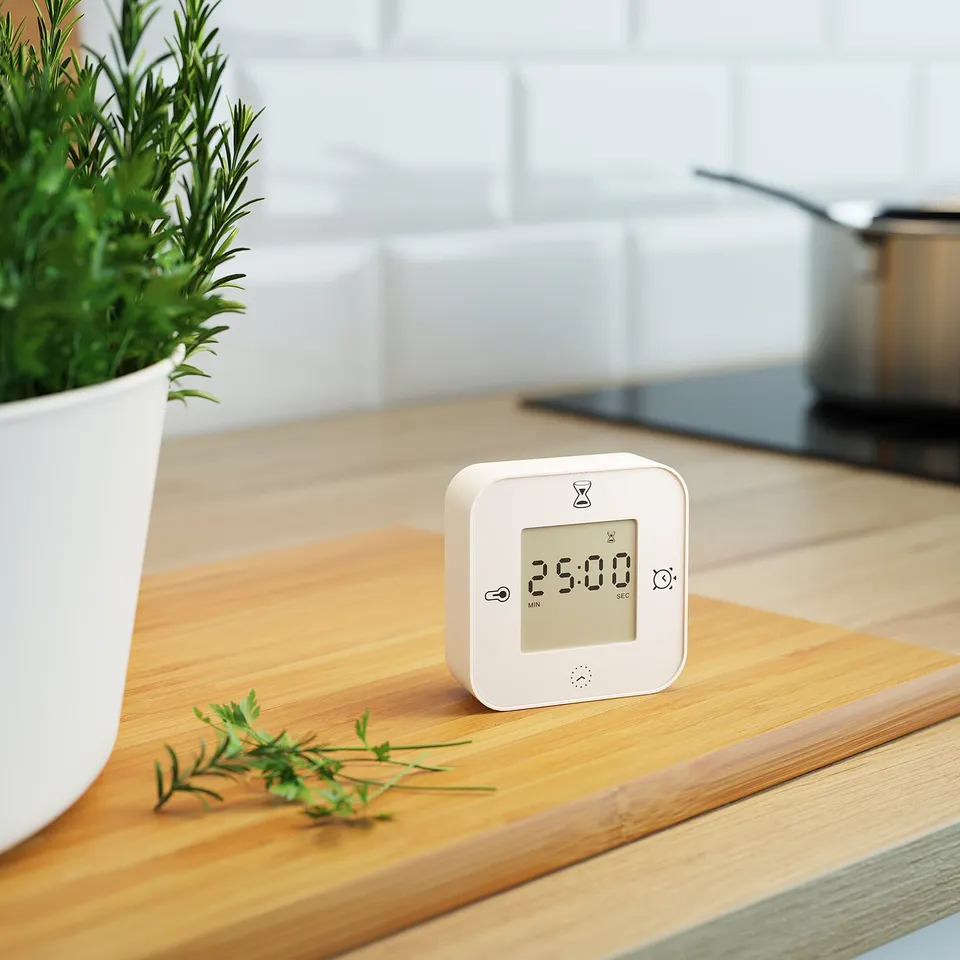 Small digital clock on wood at 25:00, with functions for time, temperature, and timer. Next to a potted plant.