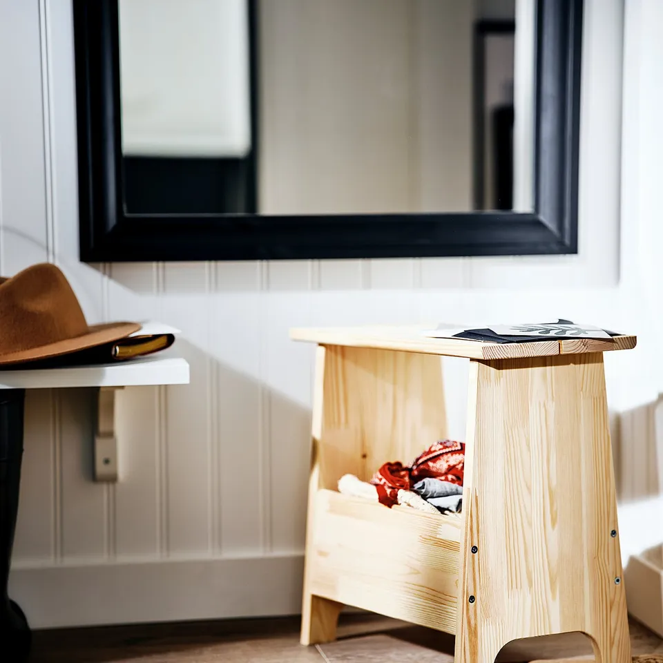 Wooden PERJOHAN stool on rug, holding clothes. Urban-chic bedroom with mirror and hat.