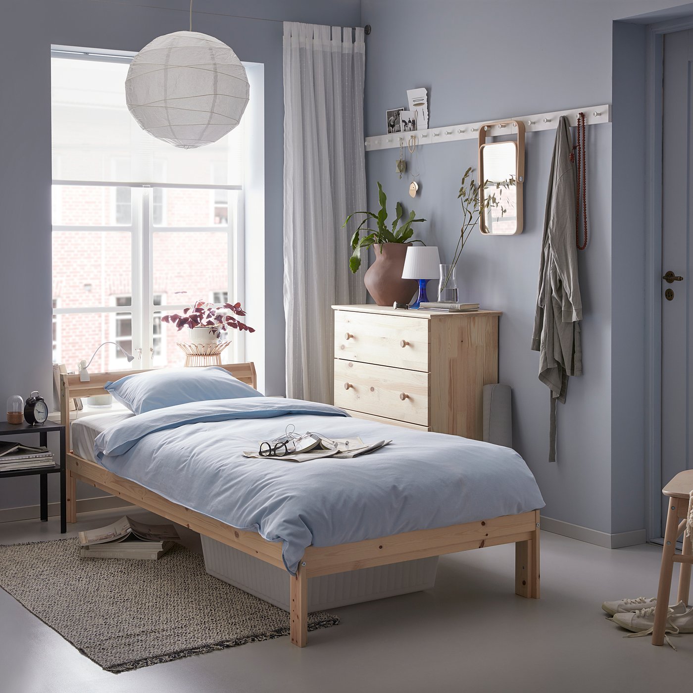 Minimalist bedroom with light blue walls, NEIDEN light wood bed, blue bedding, white pendant lamp, wooden dresser, grey curtains, and a patterned rug.