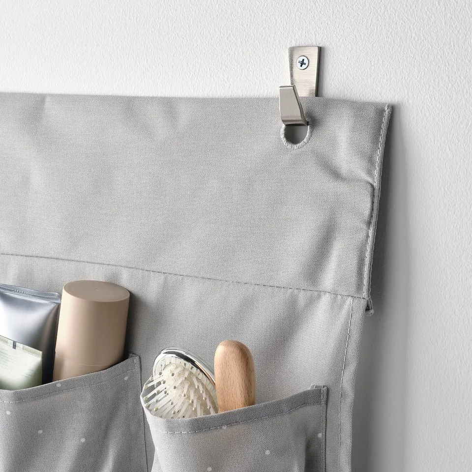 A grey hanging organiser with multiple pockets, holding a bottle, brush, and spatula. Attached to a wall with a metal hook.