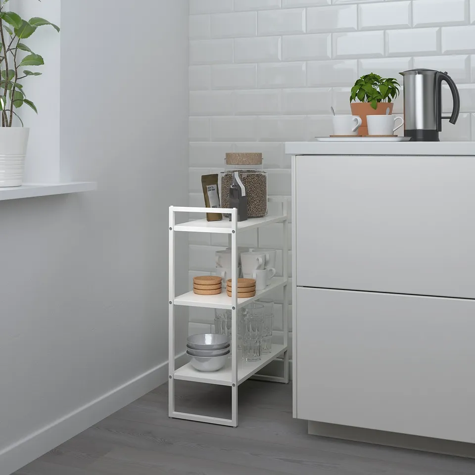 Small white JONAXEL shelf in corner with cups, bowls, and plants near larger white cabinet.