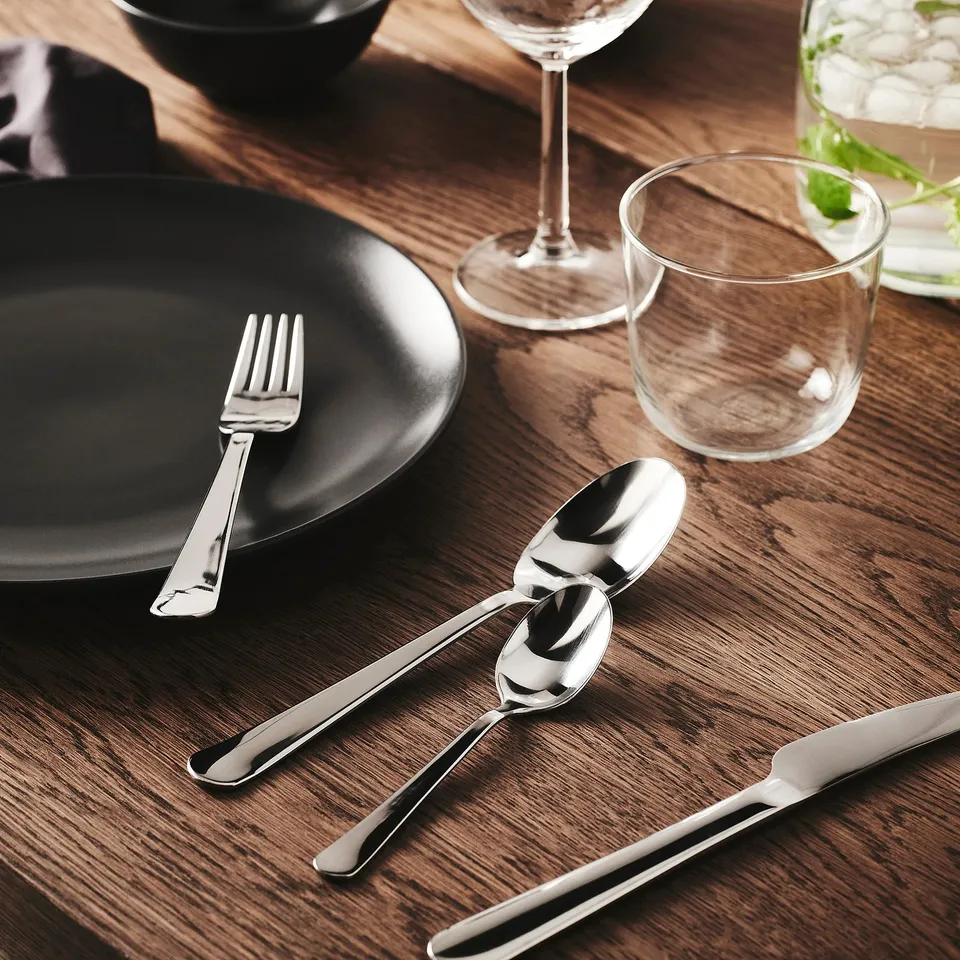 Modern dining setup with black plate, polished metal fork, knife, spoons, and glassware on a wooden table.