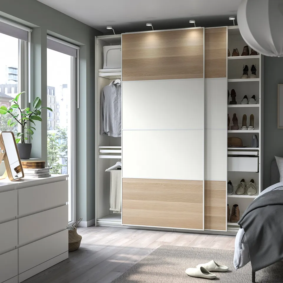 Modern bedroom with light wood PAX wardrobe, white dresser, plant, mirror, grey rug, and bed.