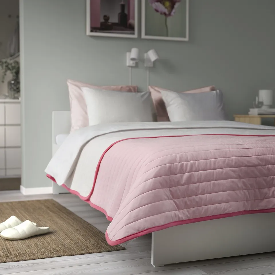 Bedroom with pink-white bedspread, grey pillows, slippers, and artwork.