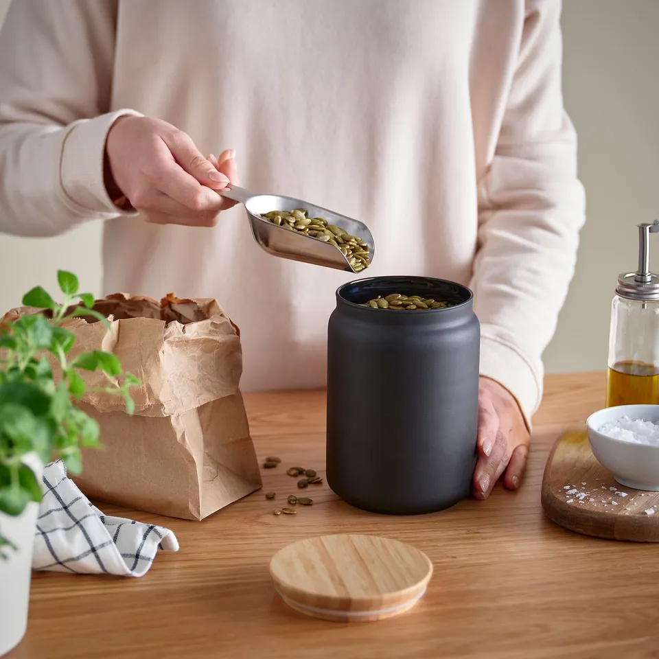 Person pours seeds into jar with lid, cloth, plant, bowl nearby.