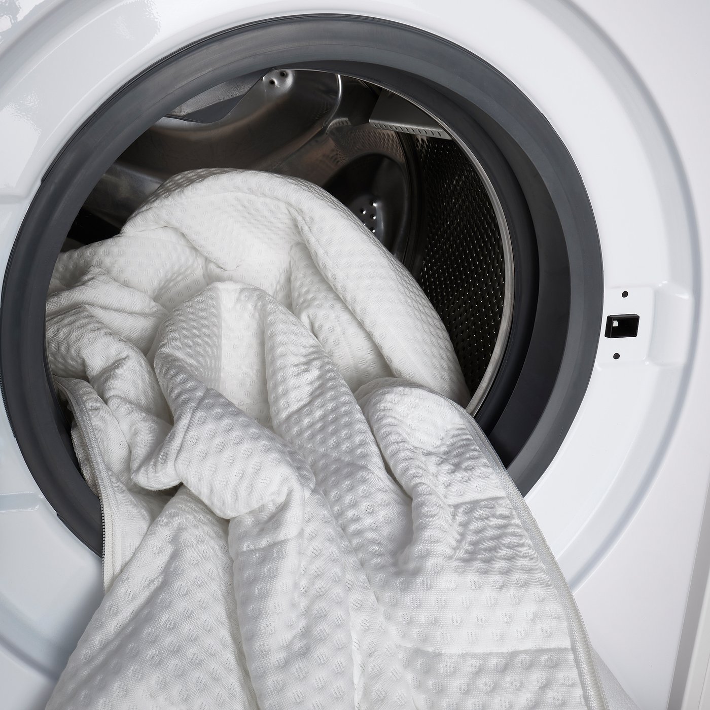 A washing machine with a white blanket inside, partially pulled out, suggesting the function of cleaning laundry.