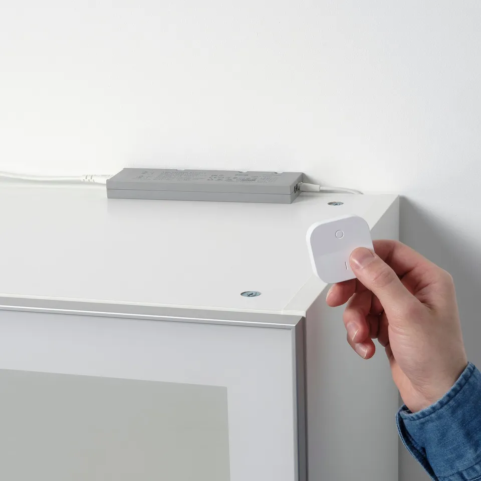 Hand pressing white remote control near a grey box on a white shelf. The box is connected by a white cord.