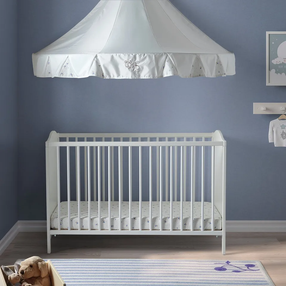A white SMÅGÖRA crib with a canopy, mattress, and teddy bear on a rug.
