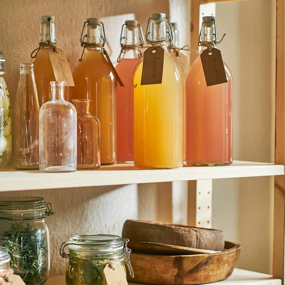 Shelf with glass bottles and jars. Bottles have varied liquids, stoppers and labels. A jar holds herbs and another jar with bread in a wooden bowl.