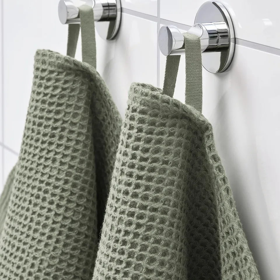 Two green VALLASÅN towels hang on shiny chrome hooks on a white wall. The towels are soft and absorbent, designed for drying hands.