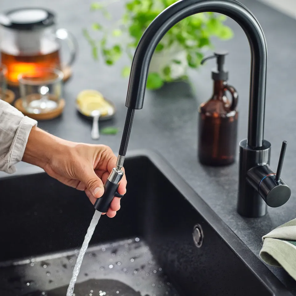 Hand holds pull-out tap spraying water into black sink. Modern kitchen setup with plants and soap dispenser.