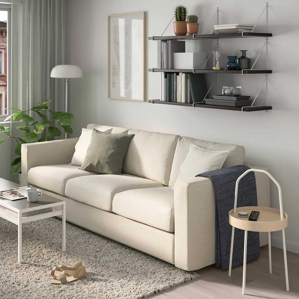 Living room with beige BERGSHULT sofa, wall shelf, side table, lamp, and plant. Light rug and curtains enhance cosy atmosphere.