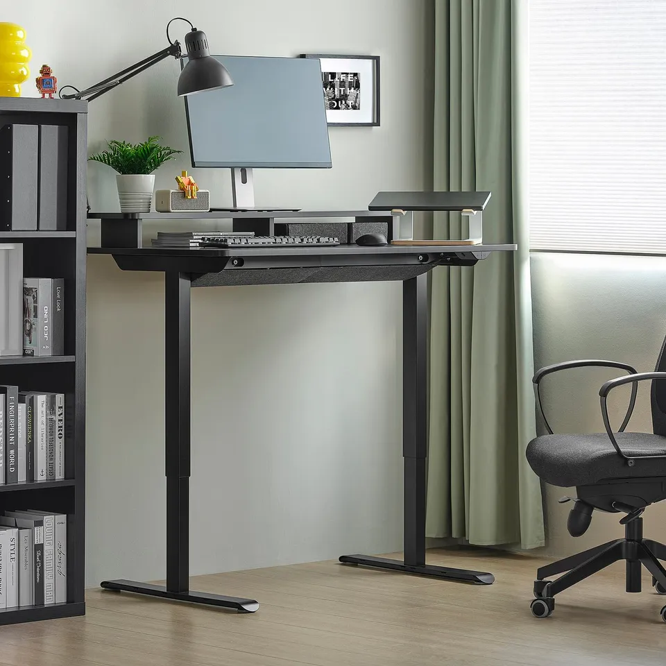 Black adjustable sit/stand desk in room with monitor, lamp, bookshelf, and office chair.