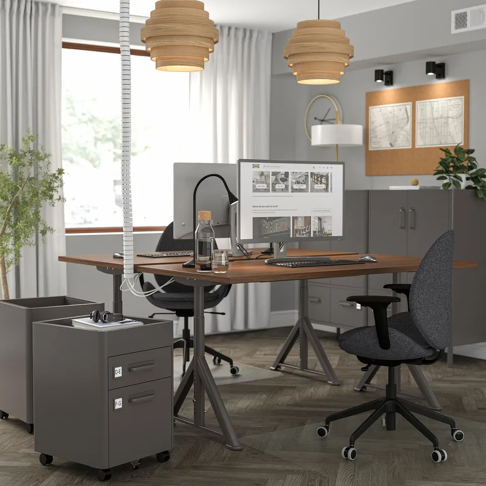 Office with IDÅSEN desks, black chairs, two monitors each. Grey cabinets, natural & pendant lighting, corkboard.