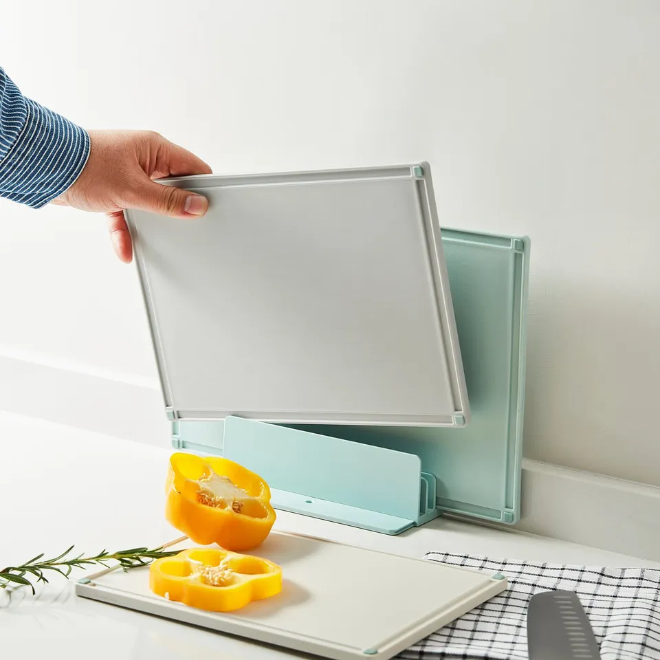 Hand holding a clear plastic chopping board with two others in holder, yellow pepper slices on counter.