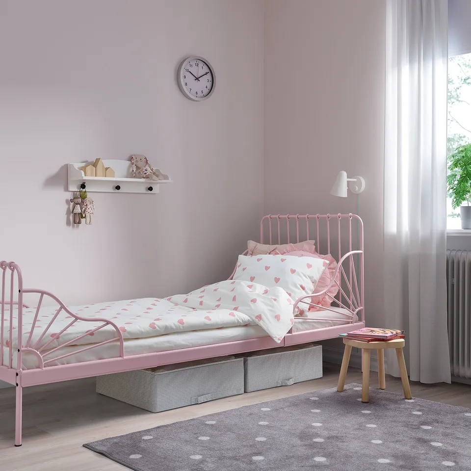 A pink MINNEN extendable metal bed in a kids room. It heart-patterned bedding and is larger than the nearby wooden stool.