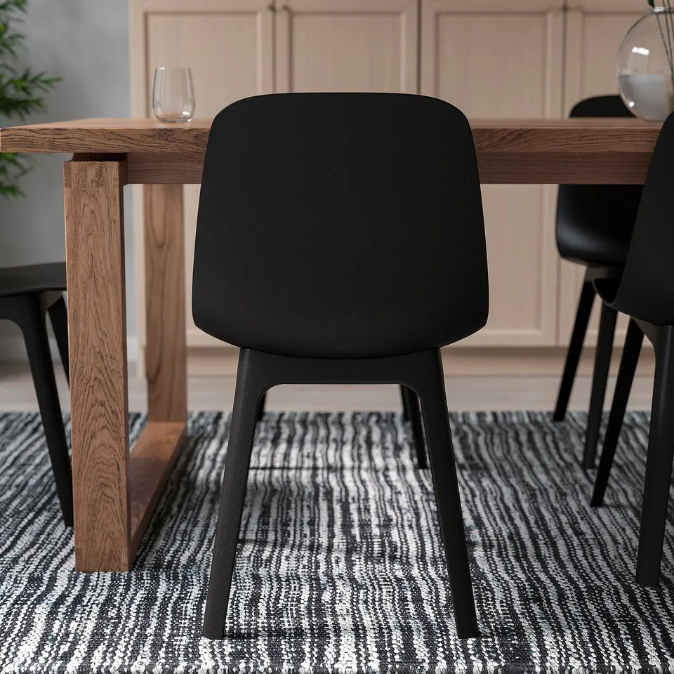 Modern wooden dining table with black chairs, one pulled out, on patterned rug, ready for easy assembly and comfort.