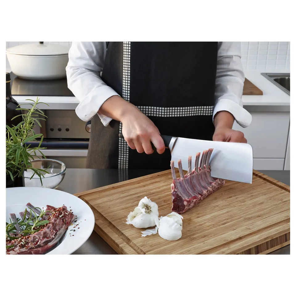 A chef uses a large, sharp knife to cut a rack of lamb on a wooden cutting board in a kitchen.
