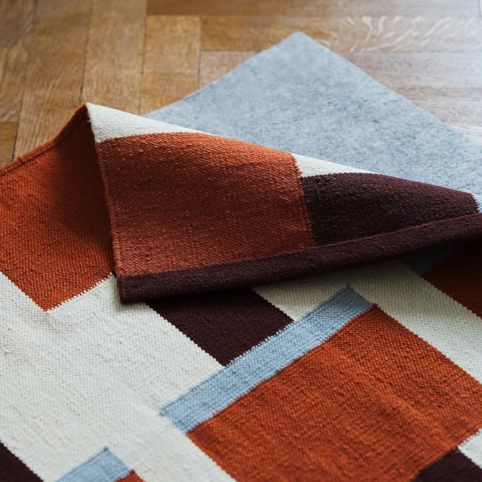 Abstract woven rug with geometric orange, white, and blue patterns laid on wooden floor. Reversible, ideal for living or dining rooms.