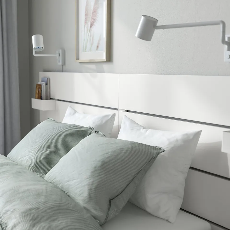Bedroom showing: NORDLI bed with drawers, headboard shelf holding books, wall-mounted lamps, white pillows, and light blue bedspread.
