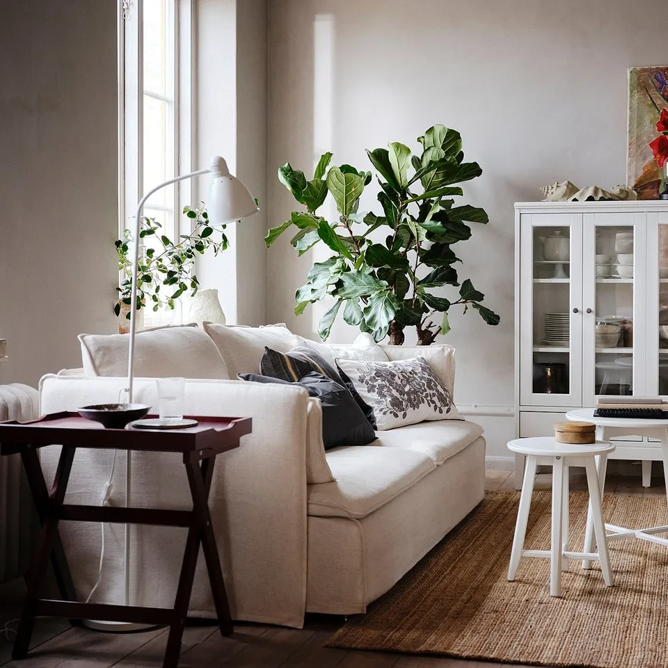 Bright living room with white sofa, wooden coffee table, jute rug, potted plant, and white cabinet with glass doors.
