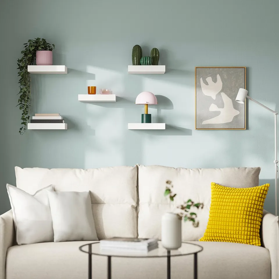 Modern living room with blue walls, white couch, colourful pillows, & minimalist white LACK shelves displaying books, vases, plants, and a lamp.