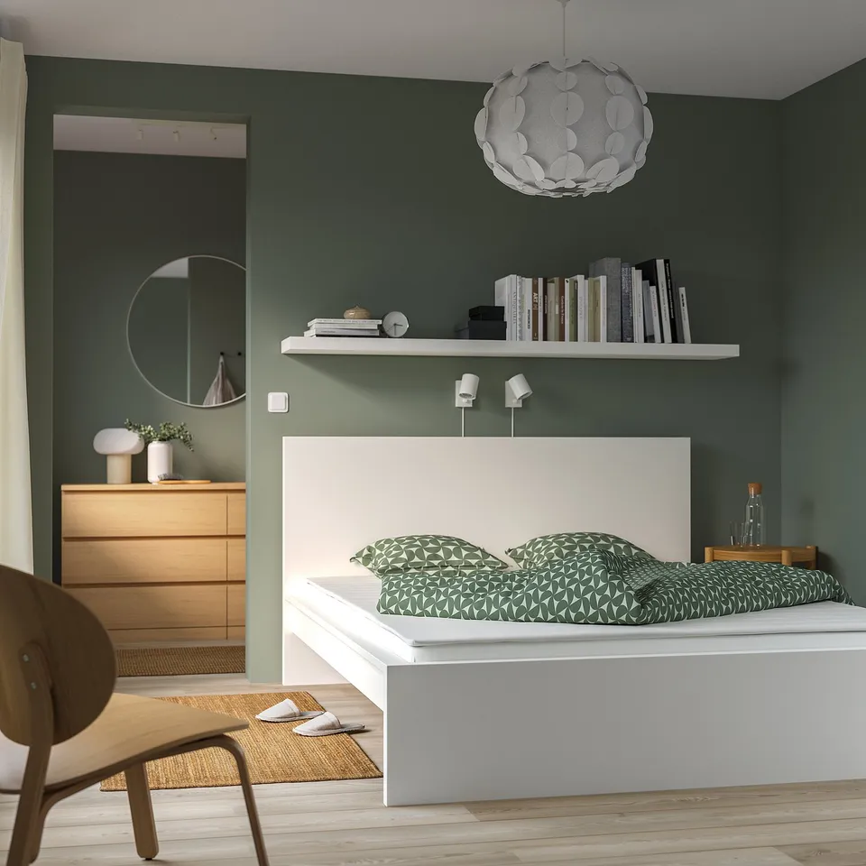 Modern bedroom with green walls, white bed, wooden dresser, unique light.