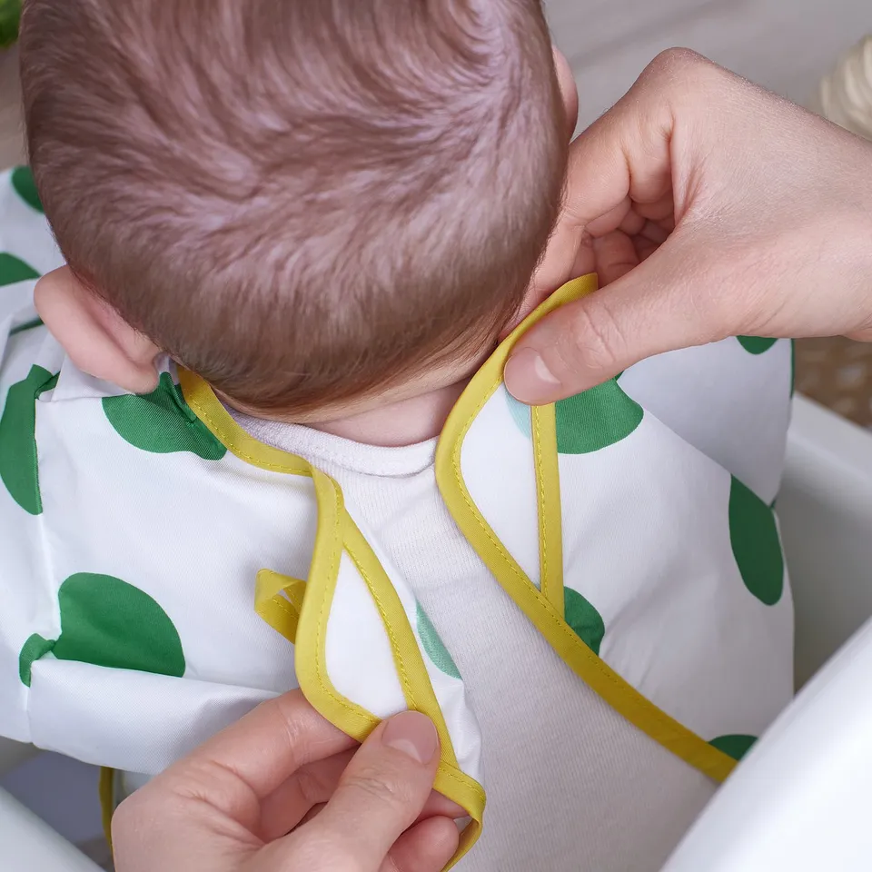 Baby wearing a white bib with green leaves being tied by an adult. The bib has long sleeves and adjustable fastenings.
