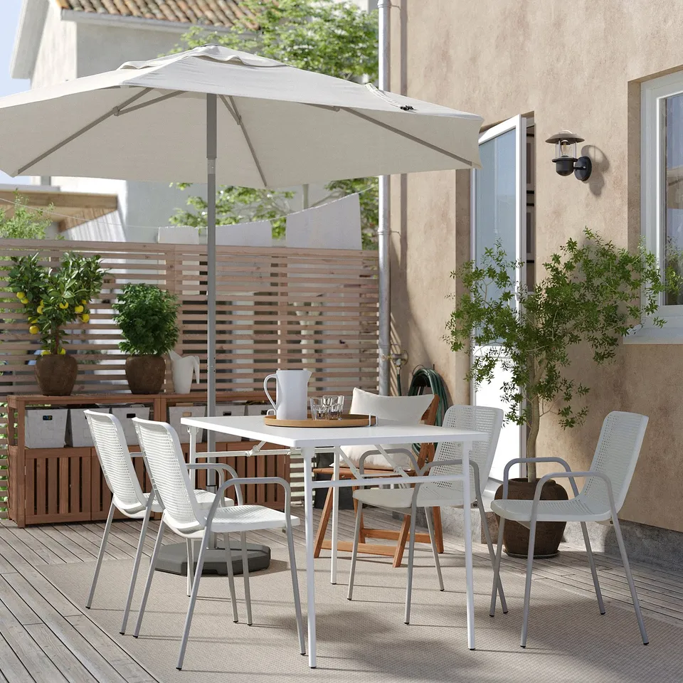 Modern outdoor dining set with white chairs and table, oversized beige umbrella, potted plants, and wooden fence; perfect for patio relaxation.