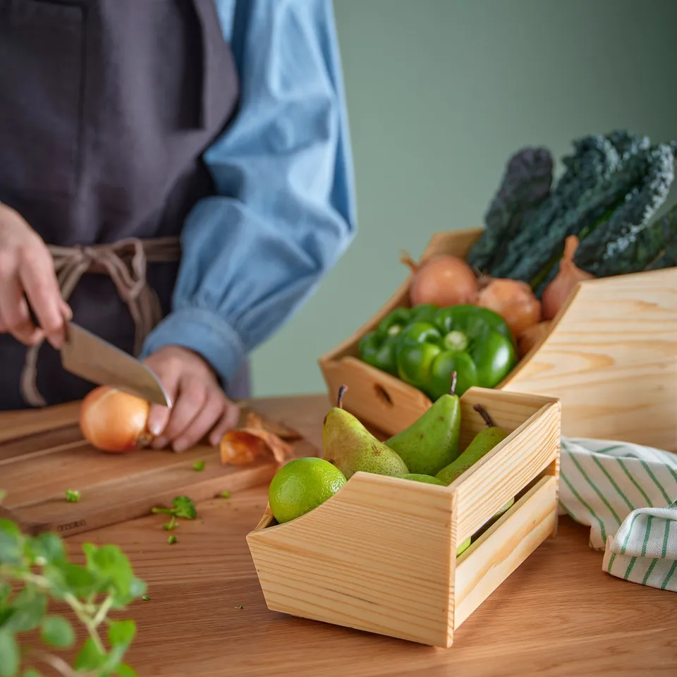 A person is cutting onions on a wooden cutting board, with other vegetables in wooden crates on a table.