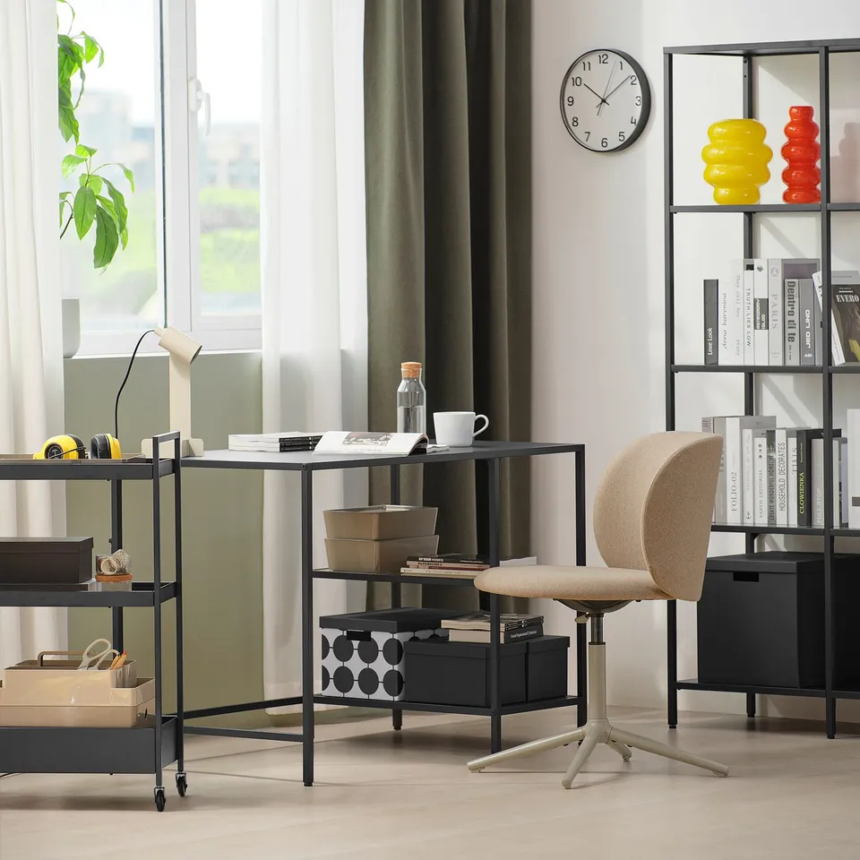 Modern home office with VITTSJÖ desk, glass top, shelves, beige chair, bookshelf with vases and books, near a window with curtains.