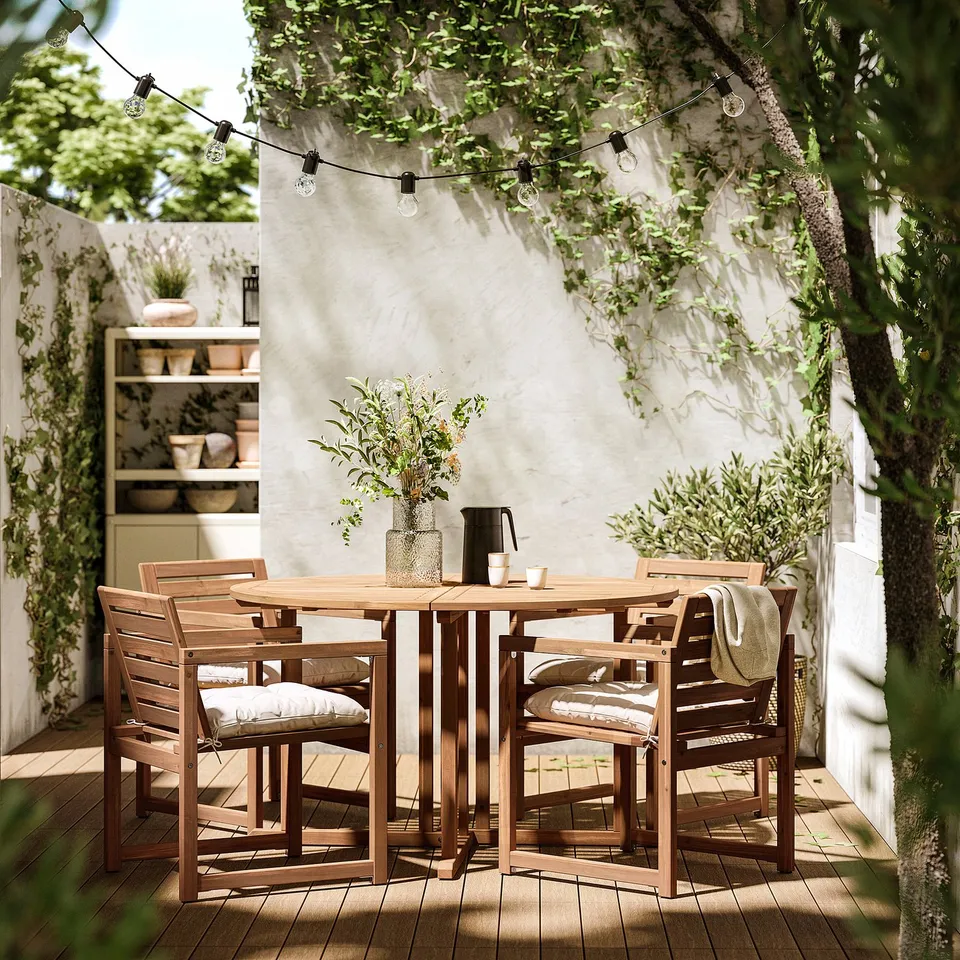 Outdoor dining set with wooden table and cushioned chairs, decorated with string lights above, set against a white wall.