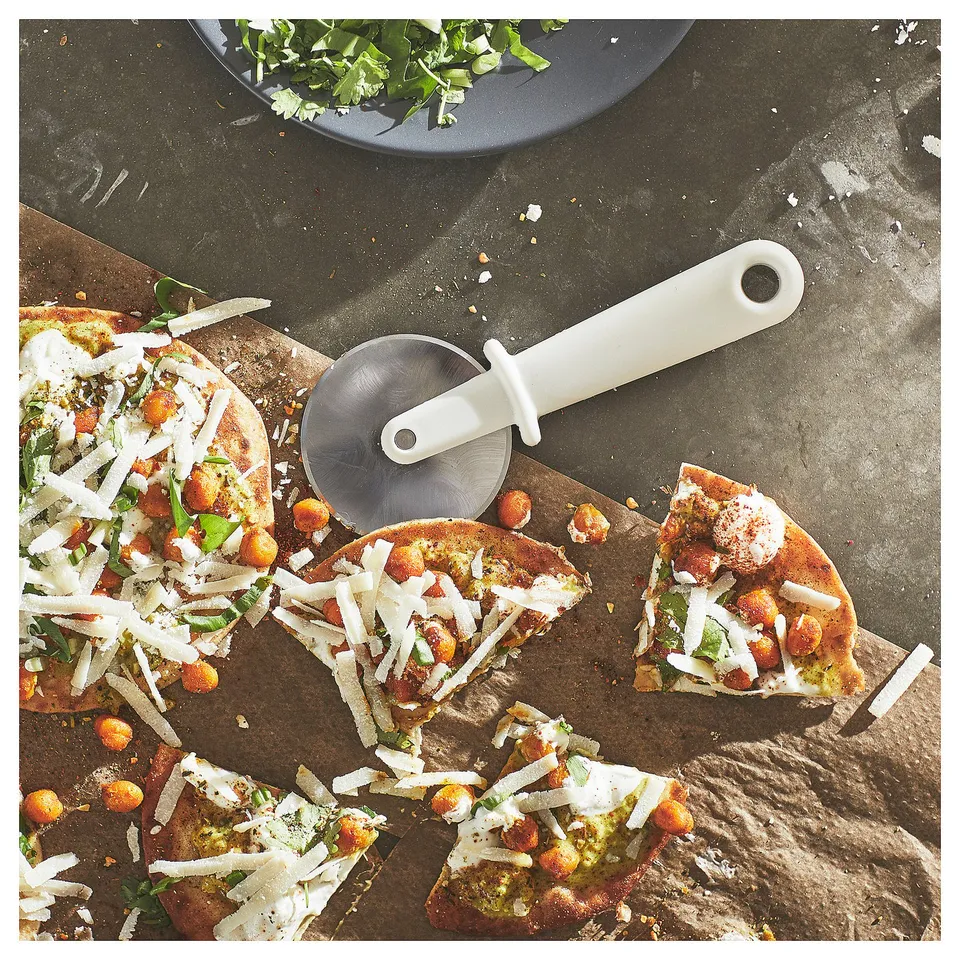 Pizza slices on stone with white-handled cutter. Topped with cheese, herbs, and tomatoes.