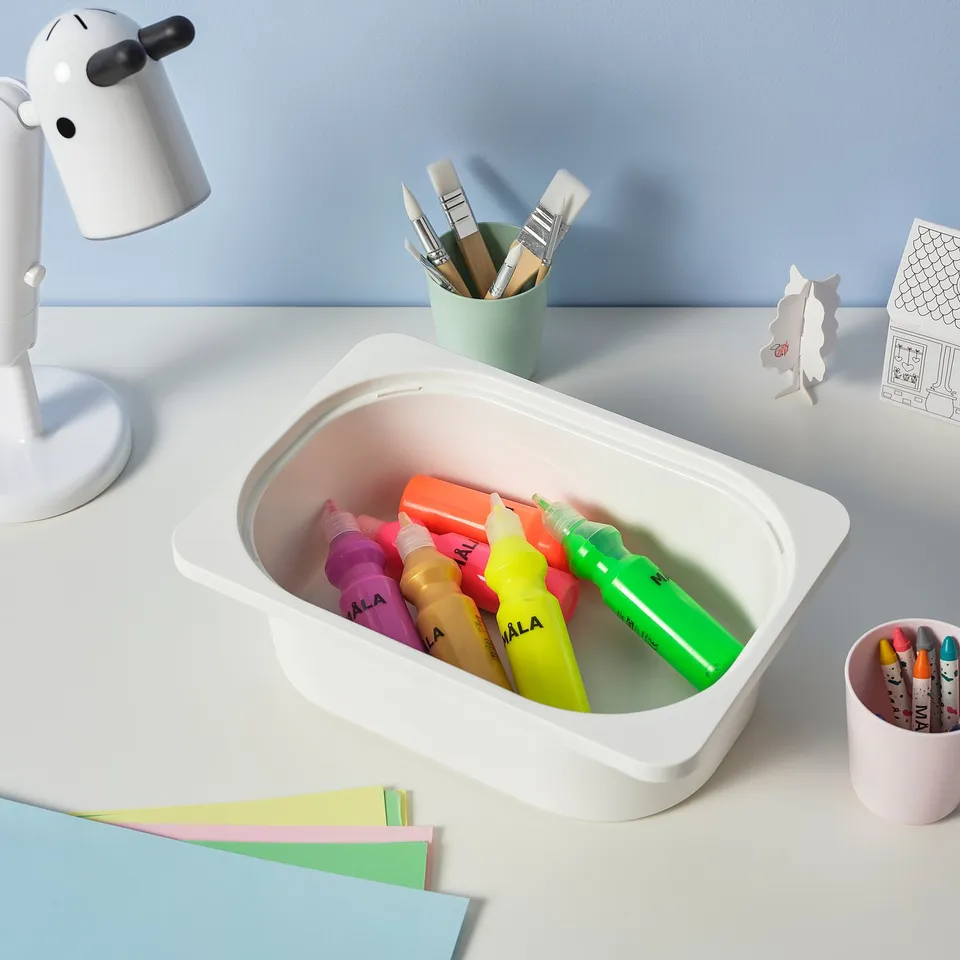 White desk with colourful markers in a TROFAST storage box, green paintbrush holder, pastel papers, and a small lamp.