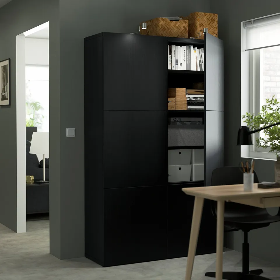Black storage unit with adjustable shelves and books. Open door reveals file folders and boxes. Desk with chair, potted plant, and lamp nearby.