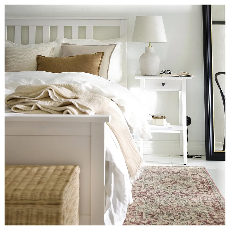 Bedroom with white bedding, beige pillows, white bedside table, lamp, and a pink, cream, and green patterned rug.