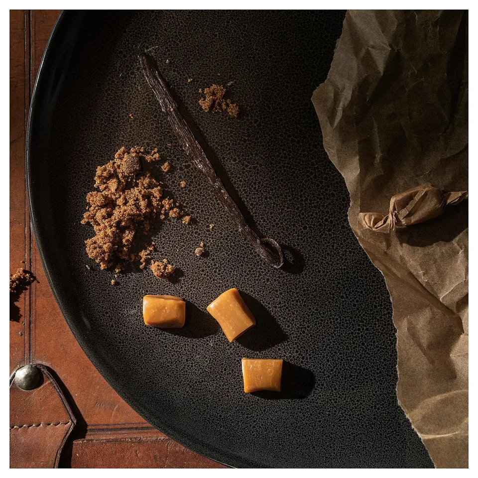 Black plate with scattered caramel candies, a vanilla bean, and crumbs. Rustic, brown paper backdrop.