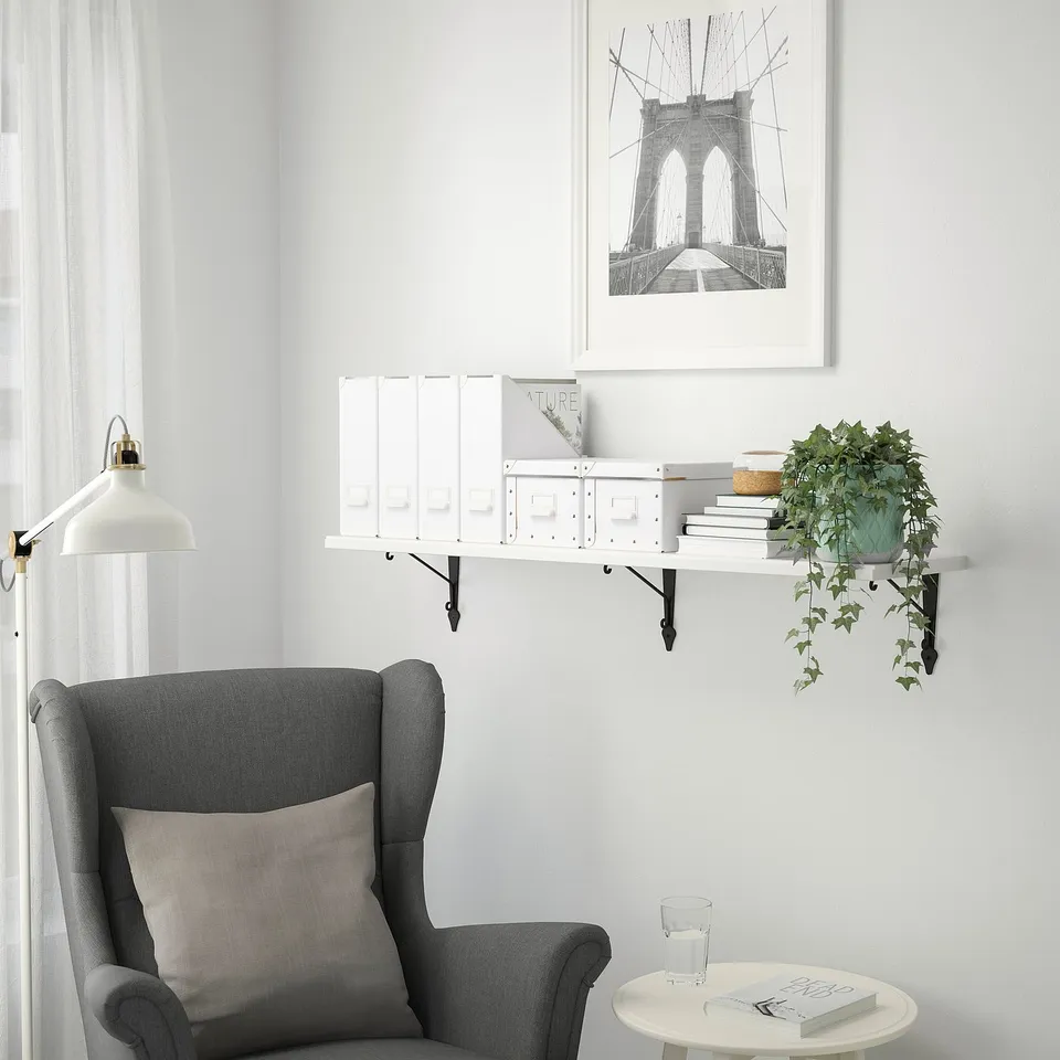 Minimalist corner with TRANHULT white wall shelf, holding books & plant; grey armchair, side table, & bridge art.