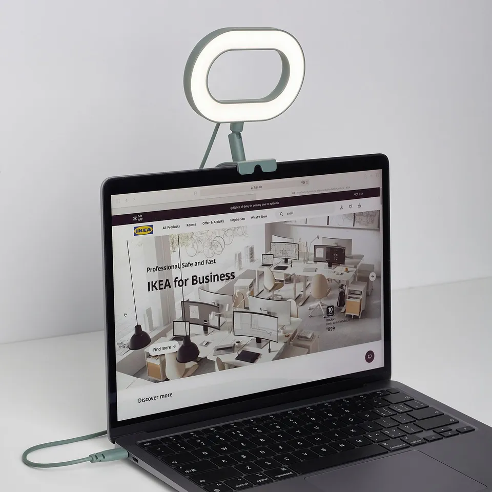 Laptop with attached ring lamp, displaying IKEA website. Lamp illuminates user for video meetings, adjustable light intensity and colour.
