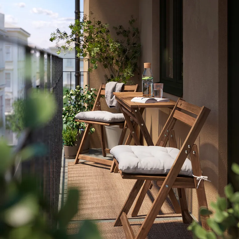Small balcony with a NÄMMARÖ brown outdoor folding table and white cushioned chairs. Glass jar and glasses on table, surrounded by green plants.