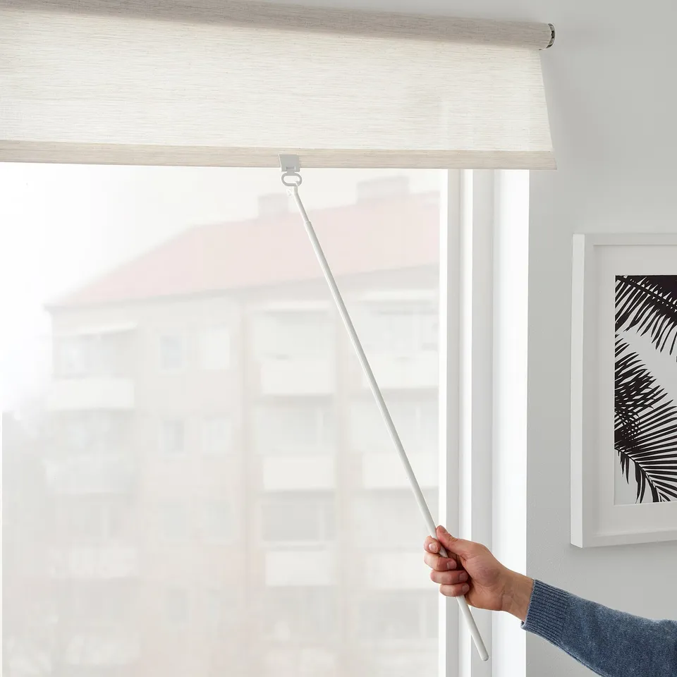 A hand lowers a light-filtering roller blind using a cordless draw rod. The blind is mounted above a window, providing privacy and reducing glare.