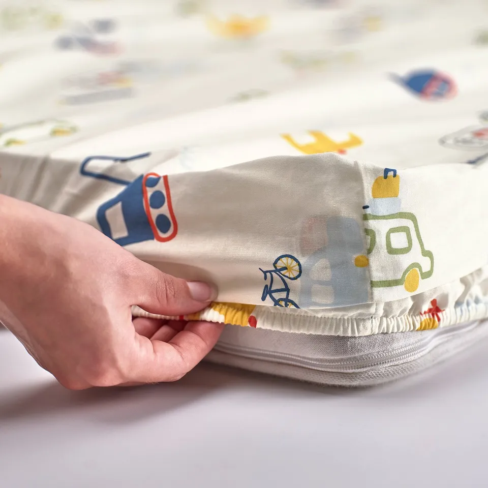A hand holding a fitted sheet with a vehicle pattern made of organic cotton, designed for a mattress up to 10 cm thick.